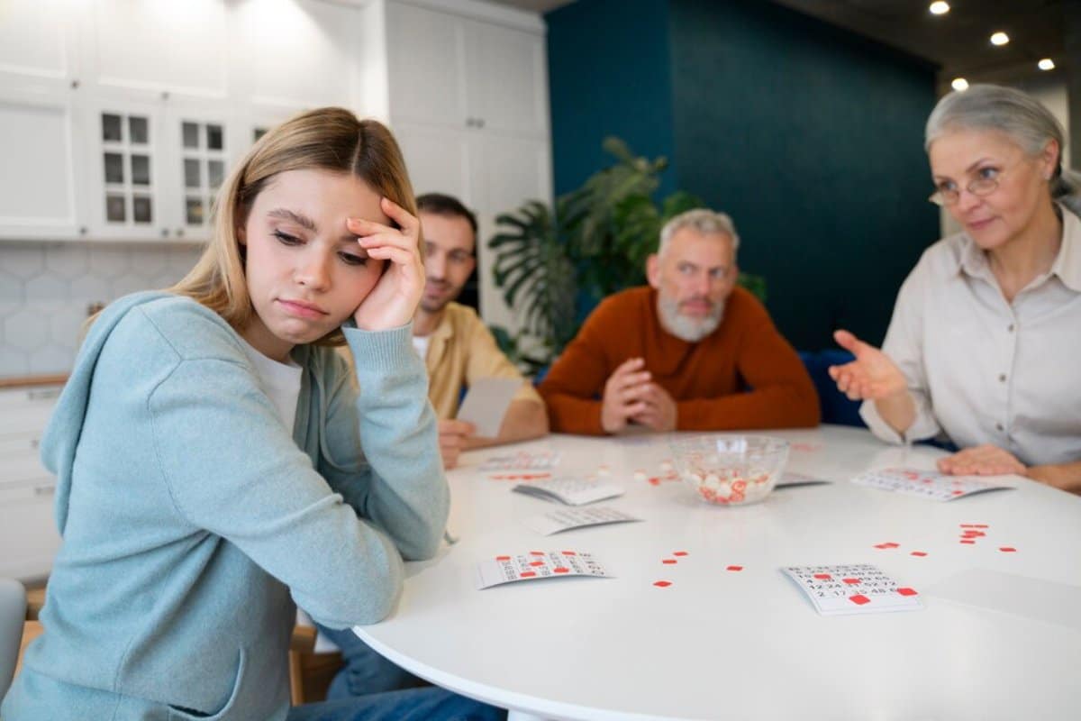 Young woman appears upset at a table while older family members discuss a card game.