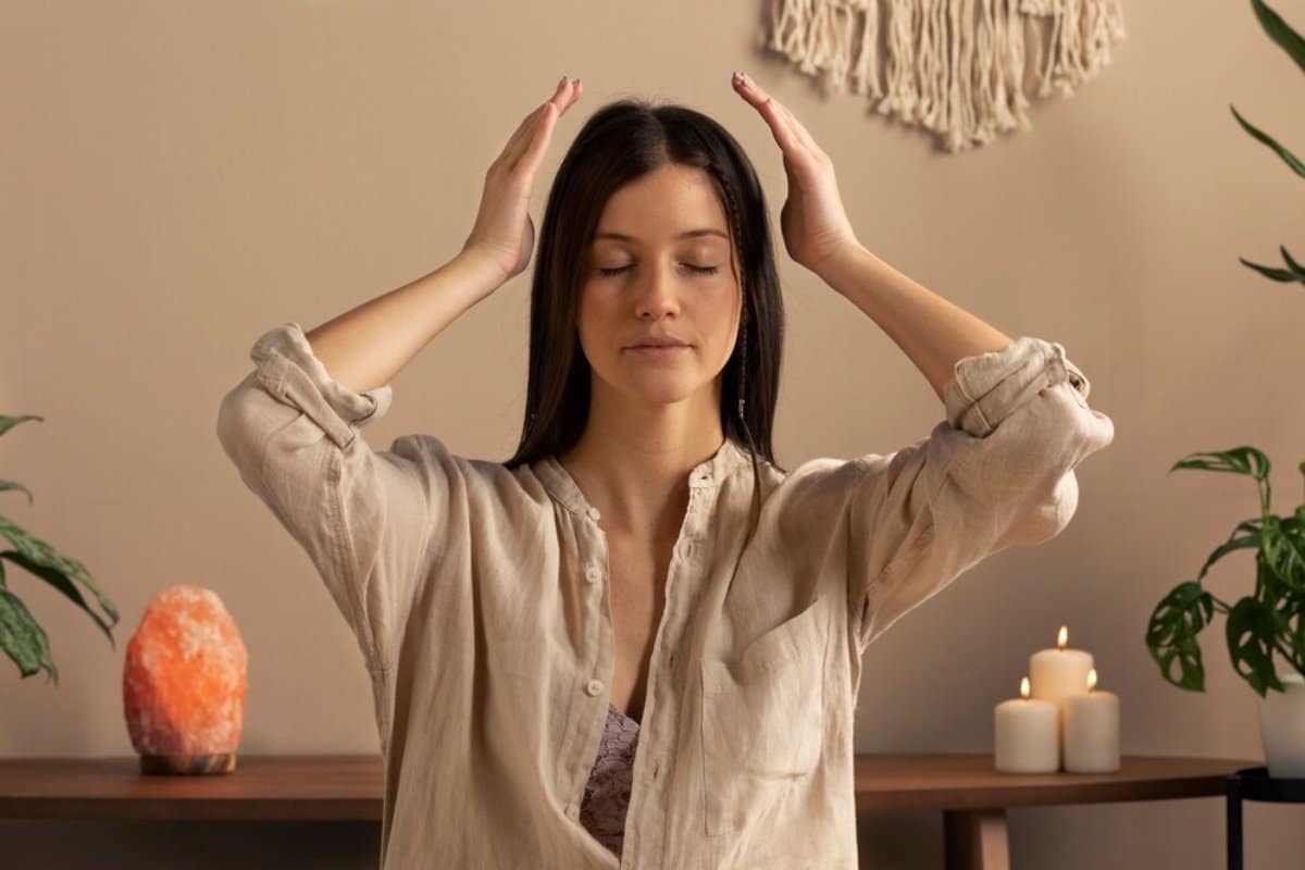 A woman meditating with her hands raised near her head, in a serene room with candles and a salt lamp.