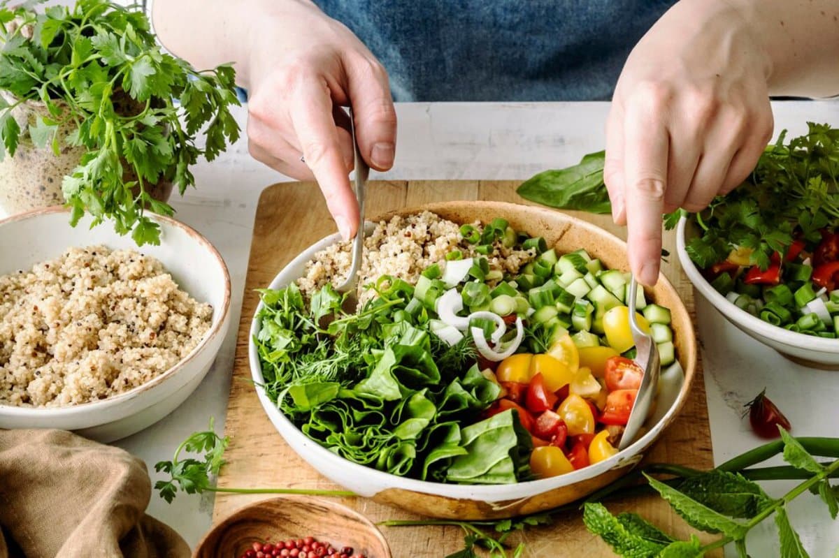 Hands preparing a fresh, vibrant salad with quinoa, cucumbers, tomatoes, onions, and assorted greens in a large bowl.