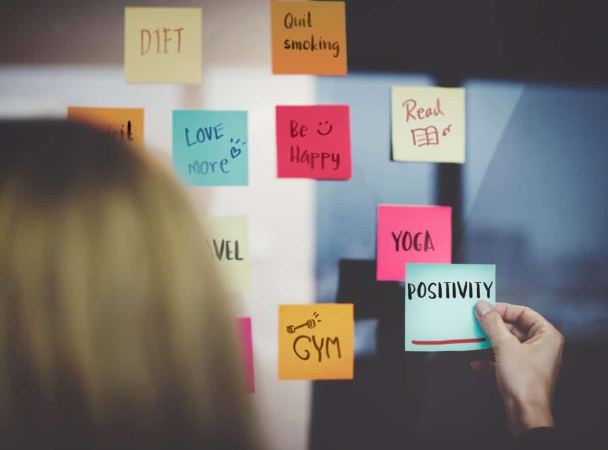 Person attaching a POSITIVITY sticky note on a board filled with various motivational reminders.