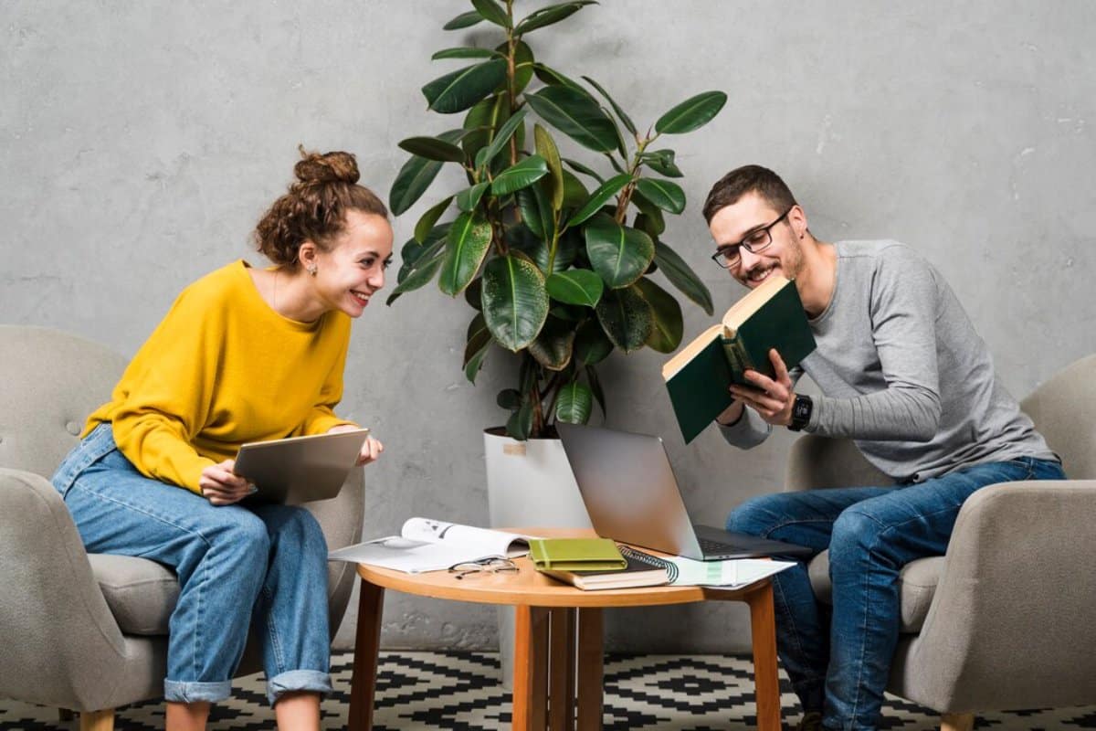 Two people smiling and engaging with digital devices and a book, seated across from each other near a large potted plant.