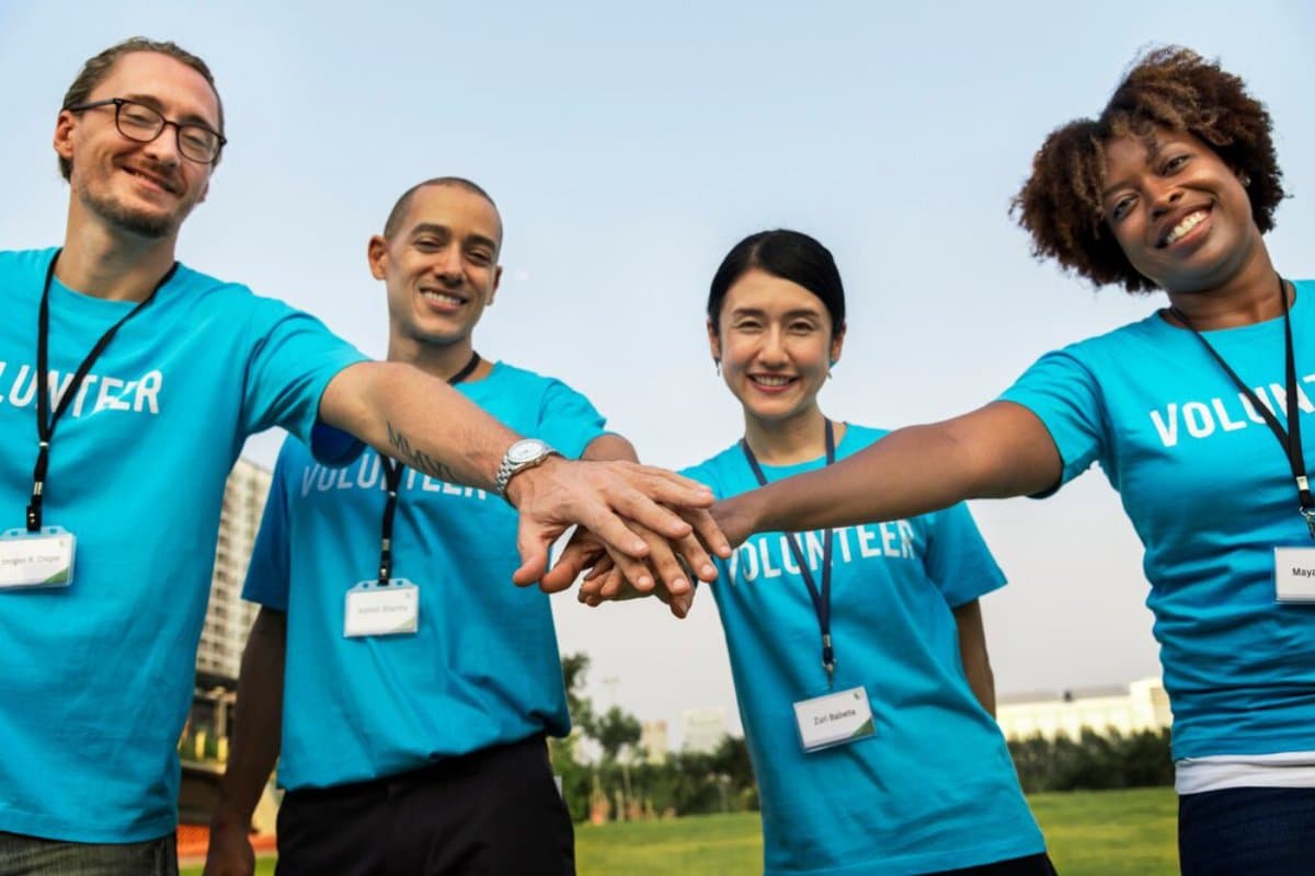 Four diverse volunteers, wearing blue 'VOLUNTEER' t-shirts, smiling and stacking hands together outdoors.