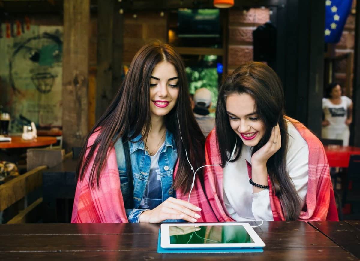Two young women sharing earphones and looking at a tablet in a cafe.