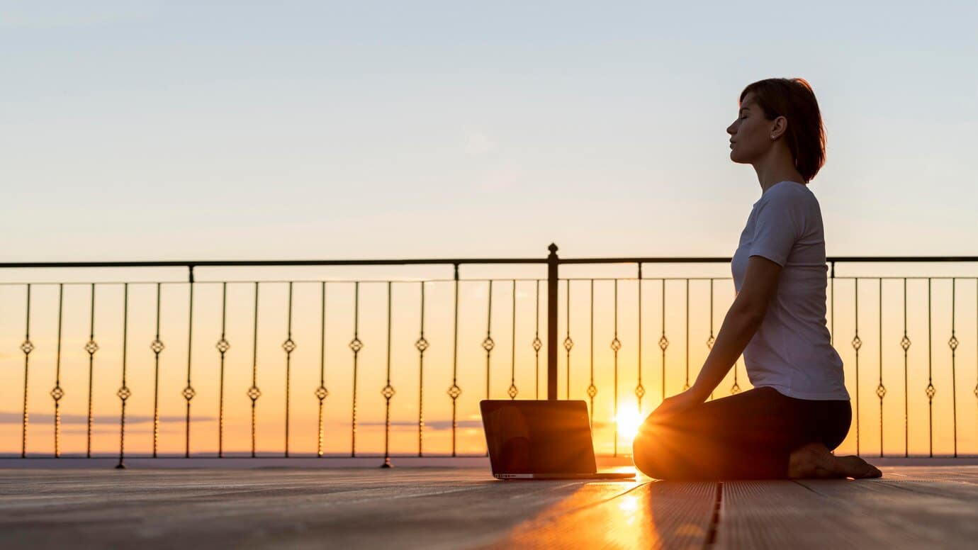 Woman meditating on wooden deck at sunset with laptop beside her.