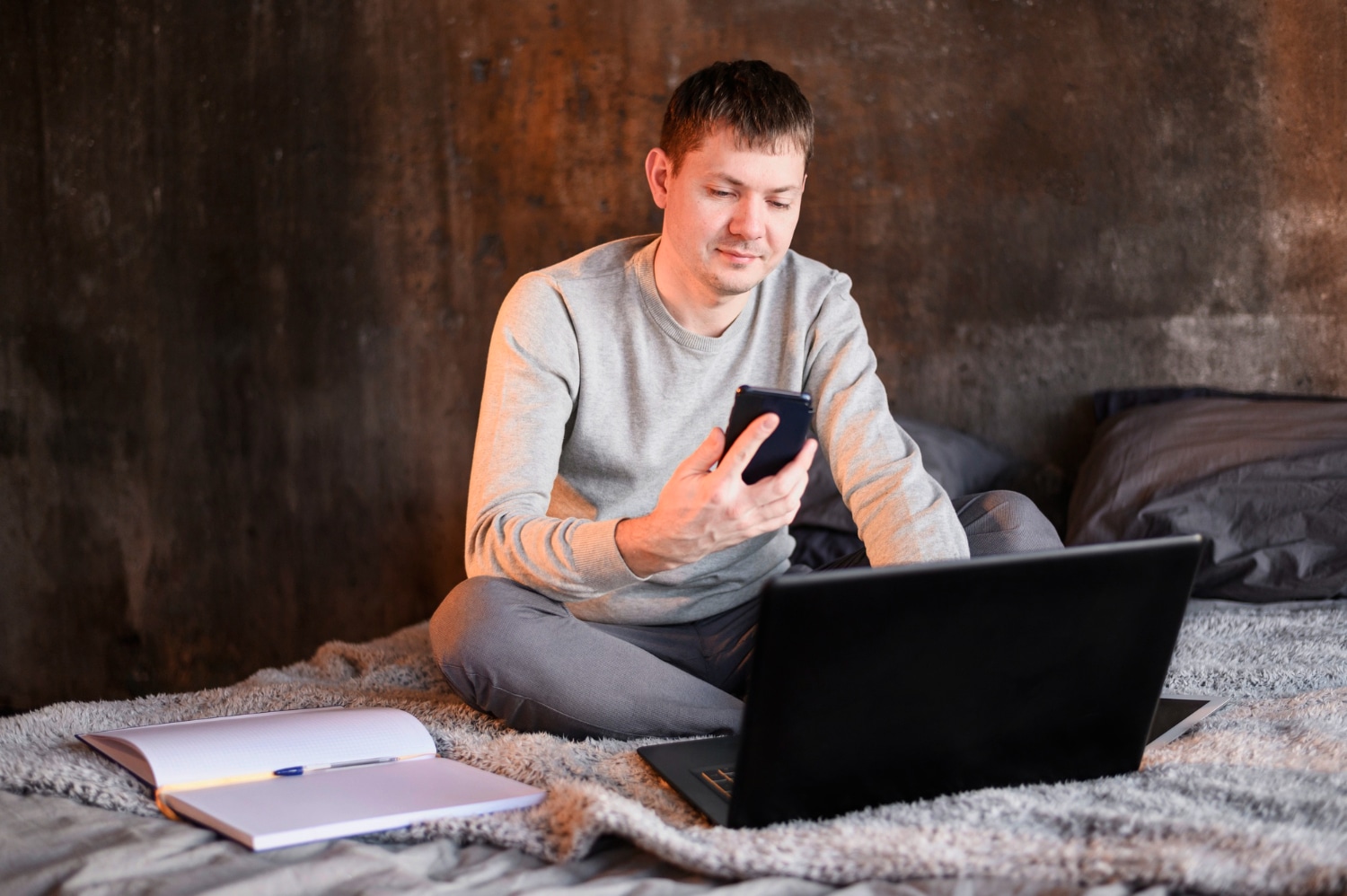 Man seated on a blanket using a smartphone and laptop, with a notebook beside him, in a dimly lit room.