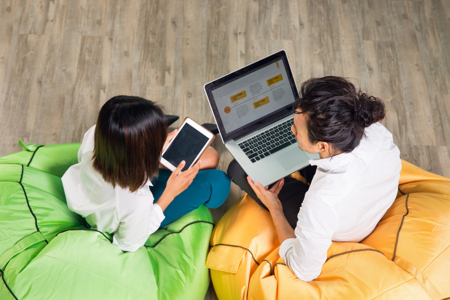 Two individuals using a laptop and tablet while sitting on colorful bean bags in a modern office setup.