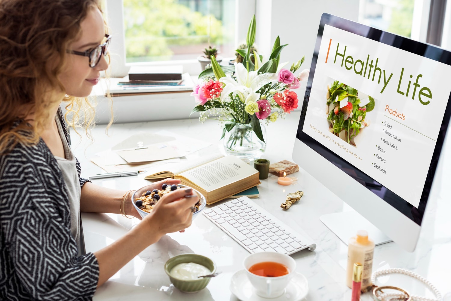Woman eating breakfast and working on a computer displaying a healthy lifestyle website, with books and flowers on the desk.