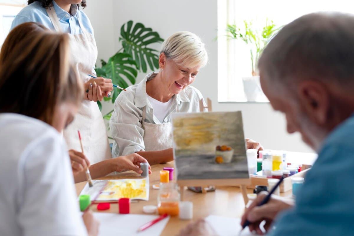 A group of older adults happily painting and discussing art in a bright, plant-filled studio.