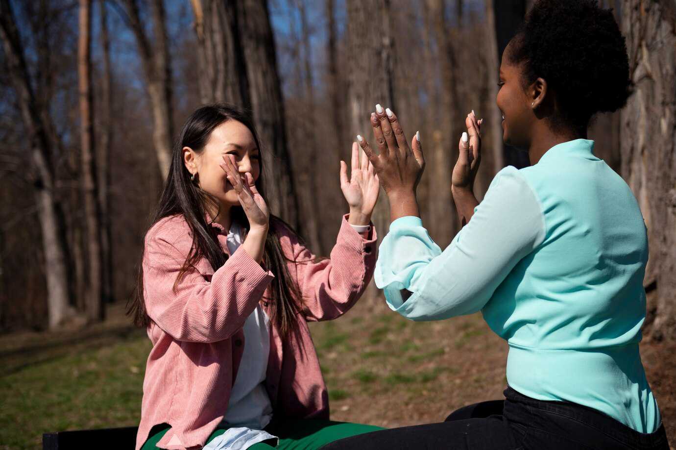 Two women face each other outdoors, engaged in gossip and playing, surrounded by tall trees in a sunny setting.