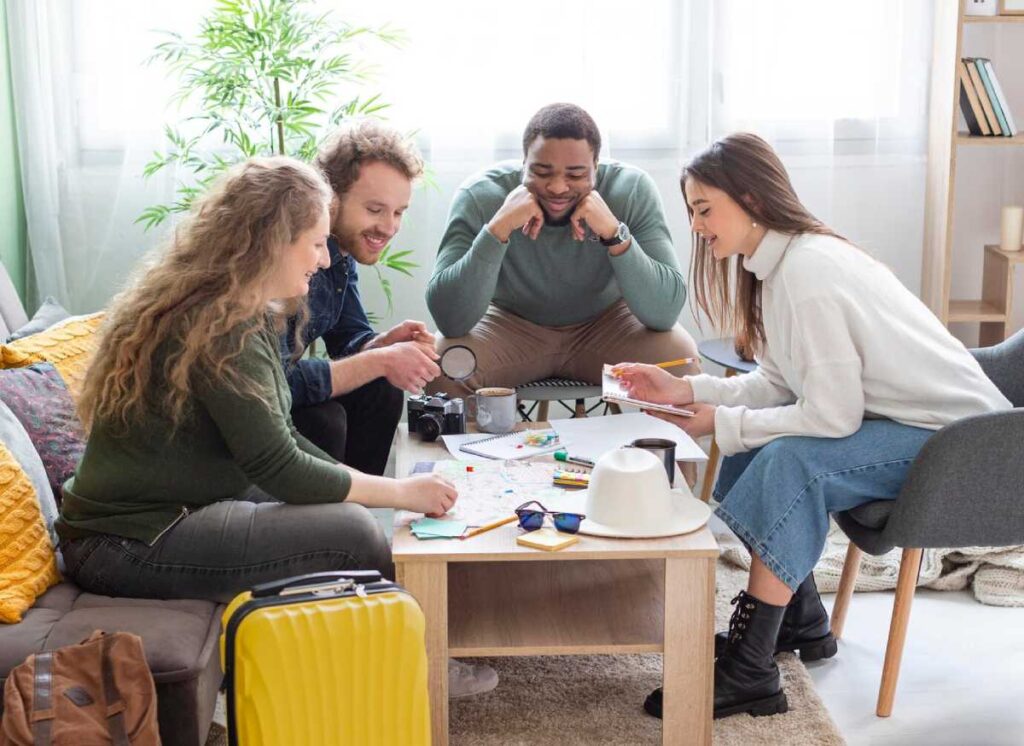 A group of four people gathers around a table with maps and notebooks, engage in activity in a bright, cozy living room.