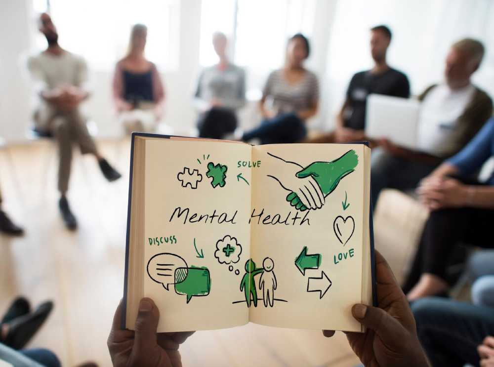 A person holds an open notebook illustrating mental health concepts with drawings of puzzles, a heart, and stick figures engaging in discussion.