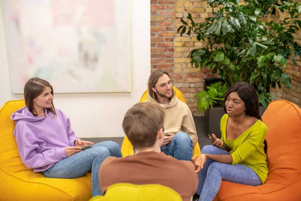A group of four friends sits on colorful bean bag chairs in a cozy, plant-filled space, engaged in conversation. 