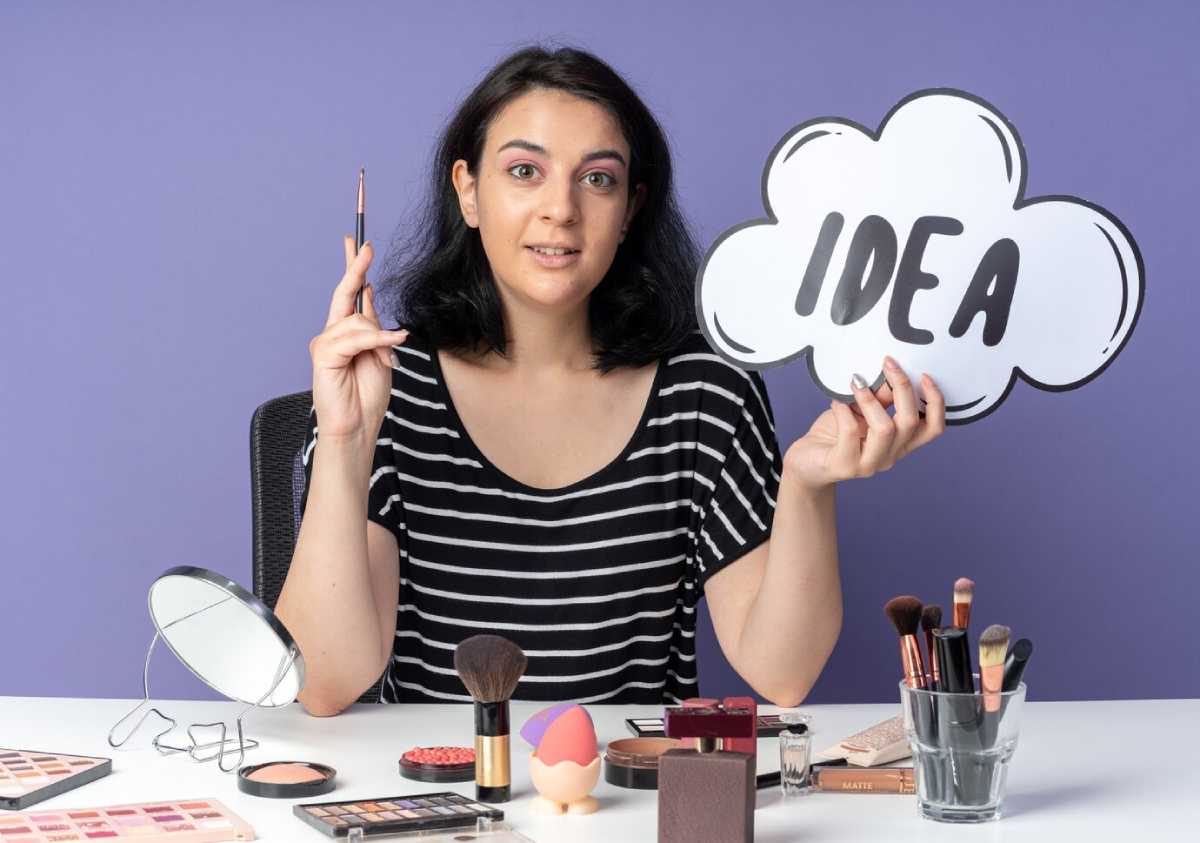A person in a striped shirt holds a sign that says "IDEA" while surrounded by various makeup products on a table.