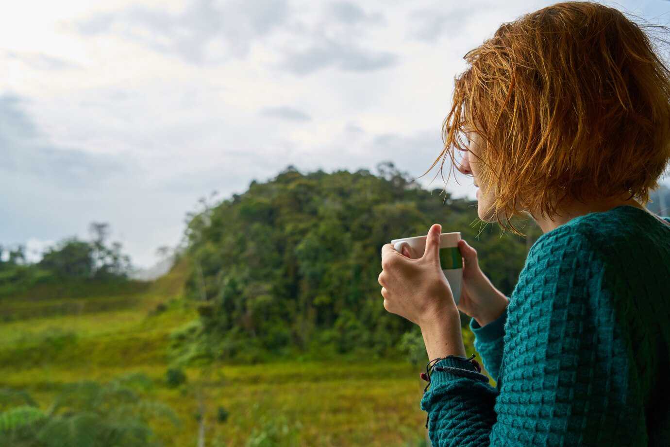 A woman admires a lush green landscape while holding a cup of coffee, under a cloudy sky, exuding a sense of calm and contemplation.